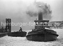 178_051 Ein Schlepper unter Dampf im Vulkanhafen - dick steigt der Qualm in den winterlichen Hamburger Himmel; das Wasser des Hamburger Hafens ist mit Treibeis bedeckt. Ein Schiffer auf einer Schute hat ein Tau in der Hand und bereitet sich darauf vor, das Schiff am Anleger festzumachen. Im Hintergrund Trmmer des U-Boot Bunkers Elbe 2; in dem 1941 fertig gestellten Bunker wurden die auf der Howaldtswerft gebauten U-Boote ausgerstet. Der Bunker hatte eine Deckenstrke von 3m - nach Kriegsende wurde die Anlage von britischen Truppen mit 47 Waggonladungen Fliegerbomben gesprengt.
