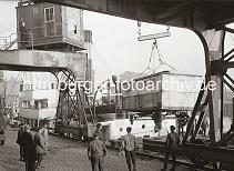 02253_579 Die Ladung eines Frachter wird vor dem Kaischuppen C des Magdeburger Hafens gelscht. Ein Zirkuswagen hngt an dem Haken eines Hafenkrans und wird an Land gebracht. Kaiarbeiter stehen mit den Hnden in den Taschen un der dem Kranportal und sehen der Arbeit zu.