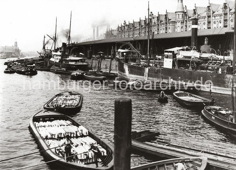 0142_233_1  Blick in den Sandtorhafen - Frachter liegen am Kai; Binnenschiffe und Schuten haben lngsseits fest gemacht. Im Vordergrund bewegen zwei Ewerfhrer ihre mit Holzkisten beladenen Schuten mit dem Peekhaken / Bootshaken durch das Hafenbecken. Links im Hintergrund ist der Kaiserspeicher zu erkennen und re. zwischen den neogotischen Giebeln der Speicherstadt am Sandtorkai die qaulmenden Schornsteine des Kesselhauses. Die Hamburger Speicherstadt wurde ab 1883 errichtet. Dabei wurden zunchst die ab dem 16. Jahrhundert auf den Elbinseln Kehrwieder und Wandrahm gewachsenen Wohnviertel abgerissen. Der Kehrwieder galt als Arbeiter- und Handwerkerviertel mit teilweise enger Gngeviertelbebauung, der Wandrahm war vor allem mit Kaufmanns- und Brgerhusern aus dem 17. und 18. Jahrhundert bebaut und insbesondere von hollndischen Einwanderern geprgt. Die Realisierung und Verwaltung der Speicherstadt wurde im Jahr 1885 durch die Hamburger Freihafen-Lagerhaus-Gesellschaft (HFLG) bernommen. Bis 1889 entstanden zwischen Kehrwiederspitze und Kannengieerort etwa 60 Prozent der Lagerflchen (Blcke A bis O). Im zweiten Bauabschnitt von 1891 bis 1897 wurden die Speicherblcke P, Q und R am St. Annenufer und Neuer Wandrahm errichtet. Im dritten Bauabschnitt von 1899 bis 1927, unterbrochen durch den Ersten Weltkrieg und die Inflationsjahre, wurde das Gebiet stlich der Strae Bei St. Annen fertig gestellt (Blcke S bis X). Das "Rathaus" der Speicherstadt, das Verwaltungsgebude der HHLA, wurde 1903 eingeweiht. Insgesamt entstanden 24 Speicherblcke mit rund 300.000 Quadratmeter Lagerflche.