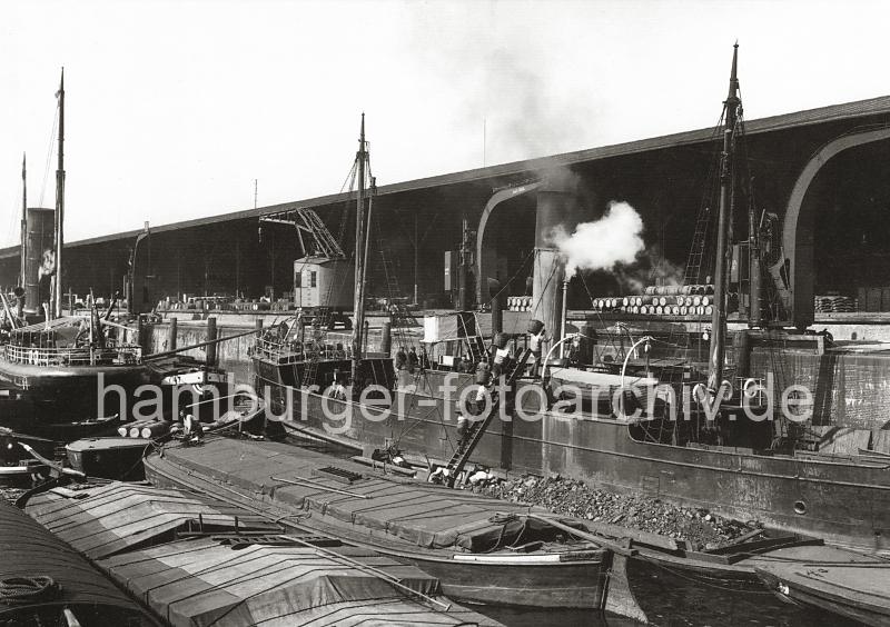 0143_230_2 Blick in den Grasbrookhafen - abgedeckte Binnenschiffe mit eigenem Steuerruder liegen im Hafenbecken des Grasbrookhafens. ber eine mit Kohle beladene Schute transportieren Arbeiter die Kohle in Krben ber eine Leiter an Deck eines Dampfschiffs. Im offenen Lagerraum des Kaischuppens sind gestapelte Fsser zu erkennen; davor laufen Dampfkrane und Rollkrane auf Schienen. 1868 wurde als erstes knstliches Hafenbecken der Sandtorhafen ausgehoben. Die neugeschaffenen Kaianlagen des nrdlichen Sandtorkais bestanden aus hlzernen Bollwerken, spter beim 1872 fertiggestellten Kaiserkai aus  Klinkern, an der die Seeschiffe direkt anlegen konnten. Sie verfgten meistens ber parallel zur Kaikante auf Schienen bewegliche Krananlagen fr den Stckgutumschlag, ber Eisenbahnanschluss sowie einfache Kaischuppen, die dem Sortieren, nicht aber dem Lagern von Waren dienten. Dies ermglichte zum ersten Mal einen direkten Warenumschlag in Bahnwaggons oder Fuhrwerke und galt seinerzeit als das modernste Umschlagsystem der Welt. Binnen weniger Jahre erfolgte der Bau weiterer Hafenbecken auf dem Groen Grasbrook, so des (1876), des Magdeburger Hafen (ab 1872) und des Brooktorhafen (um 1880) mit einem Durchlass zwischen beiden, 1881 kam der Grasbrookhafen hinzu.