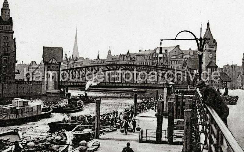 0221_222_01a Schiffsponton am Gemsemarkt bei den Deichtorhallen - Schuten mit ihrer Ladung haben am Anleger fest gemacht. Markthndler tragen ihre Ware in Kiepen zum Markt. Ein Schlepper zieht einen mit Kisten beladenen Kahn aus dem Oberhafen Richtung Zollkanal; ein weiterer Hafenschlepper fhrt gerade unter der Wandrahmsbrcke. Im Hintergrund die mehrstckigen Huser am Dovenfleet.