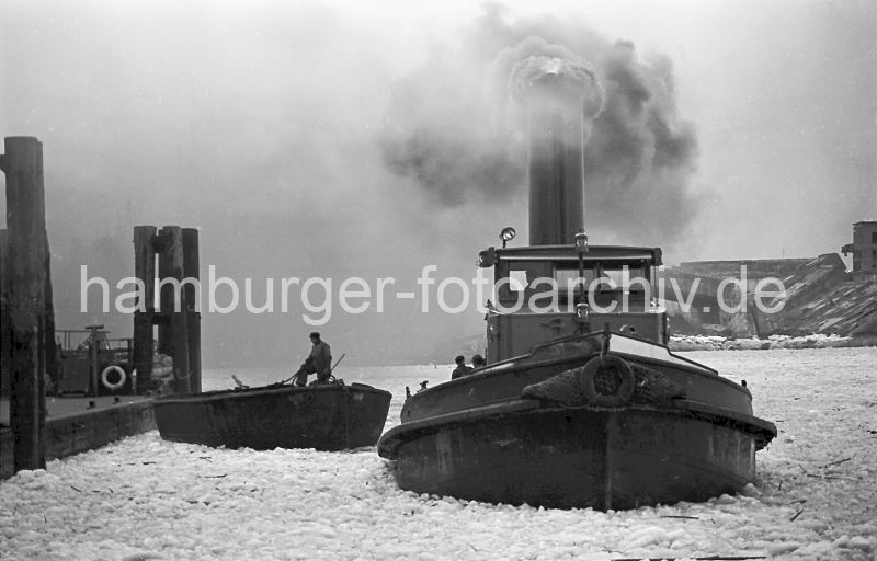 Schlepper unter Dampf - Eisgang im Vulkanhafen; Trmmer U-Boot-Bunker Elbe 2 / 1956 / 178_051 Ein Schlepper unter Dampf im Vulkanhafen - dick steigt der Qualm in den winterlichen Hamburger Himmel; das Wasser des Hamburger Hafens ist mit Treibeis bedeckt. Ein Schiffer auf einer Schute hat ein Tau in der Hand und bereitet sich darauf vor, das Schiff am Anleger festzumachen. Im Hintergrund Trmmer des U-Boot Bunkers Elbe 2; in dem 1941 fertig gestellten Bunker wurden die auf der Howaldtswerft gebauten U-Boote ausgerstet. Der Bunker hatte eine Deckenstrke von 3m - nach Kriegsende wurde die Anlage mit 47 Waggonladungen Fliegerbomben gesprengt.