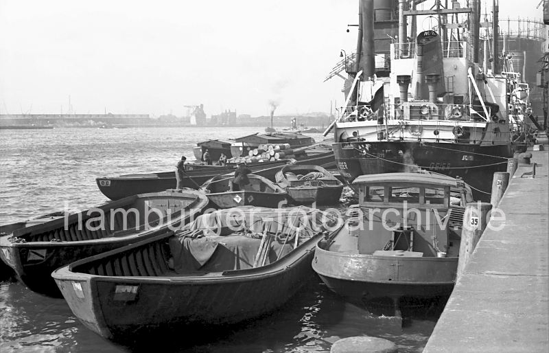 Schuten + Frachter im Baakenhafen / Versmannkai, 1956 Historische Hafenfotos aus dem Archiv der Hamburger Hafen und Logistik AG (HHLA)  182_02 Der Frachter GEIER hat am Versmannkai im Baakenhafen fest gemacht; seine Heckleinen sind um den Poller am Kai gelegt. ber Schuten, die um das Frachtschiff herum liegen, werden die Gter gelscht oder neue Fracht an Bord geladen. Auf eine der Schuten ist die Ware mit Planen abgedeckt, in einem anderen offenen Kahn liegen Fsser aufgestapelt. Zwei Ewerfhrer bringen mit Stangen ihre Schiffe in Position.  Rechts ein Schlepper, der die Khne, die keinen eigenen Antrieb haben, an ihren Bestimmungsort schleppen.