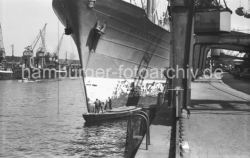 183_3 Der Frachter SCHAUENBURG liegt am Petersenkai im Baakenhafen; Matrosen des Frachtschiffs stehen auf einem Arbeitskahn und streichen den Bug des Schiffs mit Rostschutzfarbe. Im Vordergrund eine eiserne Leiter an der Kaimauer, die zum Besteigen der Khne genutzt werden kann. Daneben die Schiene, auf der die Halbportalkrane auf der Kaianlage laufen - dazwischen ein Gleis fr die Gterzge.  Frachter im Baakenhafen / Petersenkai, 1955