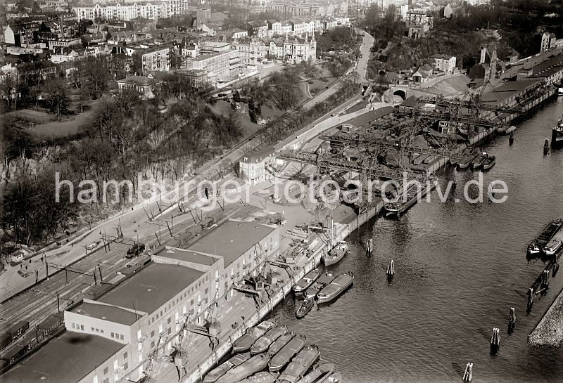 Luftaufnahme Kohlehafen Altona - Lagerhaus D / Schellfischtunnel; ca. 1932 278_3826 Krananlagen am Altonaer Kohlehafen - Binnenschiffe und Schuten liegen am Kai und werden ent- und beladen. Im Vordergrund das Lagerhaus D am Neumhlen Kai; an der Strassenseite des Lagergebudes mehrere Eisenbahngleise mit Gterzgen und einer Rangierlokomotive. Im Hintergrund die Brcke am Elbberg, direkt lks. dahinter die Einfahrt zum Tunnel der sogen. Schellfischbahn, der Altonaer Hafenbahn, dessen Strecke zum Altonaer Bahnhof fhrt.