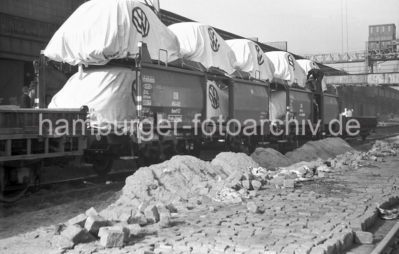 435_11 Autotransport auf Gterwaggons in den Hamburger Hafen; die Volkswagen stehen zum Schutz mit Planen auf Waggons auf einer Kaianlage im Oderhafen, einem der zahlreichen Hafenbecken im Hamburger Hafengebiet. Im Hintergrund der Ausleger einer Krananlage, die sich ber den Bahngleisen befindet. Bauarbeiten auf dem Hafenkai, die Strasse zwischen den Gleisen wird mit Pflastersteinen neu gepflastert.  historische Hafenfotos aus dem Archiv der Hamburger Hafen und Logistik AG (HHLA) Gterwaggons fr den Autotransport - Stoffabdeckung VW, Volkswagen Logo 