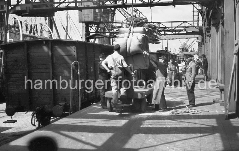 Lschen eines Frachters im Hamburger Rohafen - Gterwaggons am Kaischuppen. 439_04 Arbeit im Hamburger Hafen ca. 1948; ein Frachter wird im Rohafen gelscht. Der Kran bringt die Scke aus dem Frachtraum des Schiffs an Land - Kaiarbeiter nehmen die Ladung in Empfang. Auf einem Elektrokarren werden die Scke in den Lagerschuppen gebracht. Im Hintergrund arbeiten weitere Krane und bringen Scke an Land - Hafenarbeiter transportieren das Sackgut mit Sackkarren. Gterwaggons stehen auf der Gleisanlage direkt an der Laderampe der Lagerschuppen.