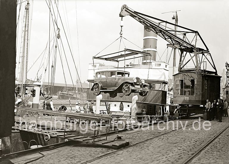 Auto - Verschiffung im Hafen Hamburg; Kran am Grasbrookhafen, 1934  440_08 Verladung eines Autos am Hbenerkai des Grasbrookhafens -  die Hafenarbeiter stehen neben dem Kran und an Deck des Frachtschiff; sie beobachten, wie das Kraftfahrzeug an Bord des Frachter gehievt wird. Mit einem speziellen Anschlaggeschirr, das um die Reifen des Wagen gelegt wird kann das Fahrzeug sicher angehoben werden Der Kran, der auf Schienen entlang der Kaianlage bewegt werden kann, stammt vom Hamburger Hersteller Nagel & Kamp / Kampnagel.