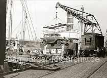 440_08 Verladung eines Autos am Hbenerkai des Grasbrookhafens -  die Hafenarbeiter stehen neben dem Kran und an Deck des Frachtschiff; sie beobachten, wie das Kraftfahrzeug an Bord des Frachter gehievt wird. Mit einem speziellen Anschlaggeschirr, das um die Reifen des Wagen gelegt wird kann das Fahrzeug sicher angehoben werden. Der Kran, der auf Schienen entlang der Kaianlage bewegt werden kann, stammt vom Hamburger Hersteller Nagel & Kamp / Kampnagel.