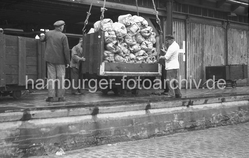 Bananen Ladung lschen;  Hieve mit Bananenbschel, Segelschiffhafen - Asiakai; 1949 446_4 Die verpackten Bananenbschel werden in einer Transportkiste von Bord des Khlfrachters an Land gehievt. Auf der Kaianlage nehmen Lagerarbeiter die Hieve mit den Bananen in Empfang - die Kiste wird auf Rollen gestellt und danach in den Lagerschuppen geschoben. Links steht eine leere Transportkiste, die danach an den Haken des Krans befestigt wird.