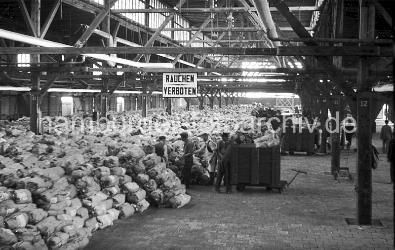 historische Hafenmotive aus dem Archiv der Hamburger Hafen und Logistik AG (HHLA)  Bananenschuppen - Lagerung, Lagerarbeiter - Transportkisten /  Asiakai 1949 448_6 Die Bananen werden im Lagerschuppen von hlzernen Transportkisten geladen und in mehreren Schichten in dem riesigen Hafenschuppen gelagert. Der Fussboden des Gebudes ist mit Steinen gemauert - eine massive Holzkonstruktione / Holzstnder tragen das Dach; oben sorgt eine Fensterreihe fr ausreichendes Licht. Fr die Arbeiten in der Nacht hngen Lampen an den Dachbalken. 
