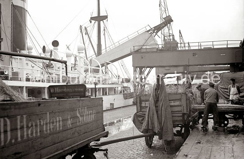 Bananen - Verladung; Pferdefuhrwerke an der Laderampe - Bananenfrachter am Kai; ca. 1930 - historische Hafenfotos aus dem Archiv der HHLA. historische Bilder aus dem Hamburger Hafen - Drucke auf Fotoleinwand / Posterprint als Geschenk fuers Jublaeum + Geburtstag. 452_10_B_317a  Hafenarbeiter haben Bananen aus dem Kaischuppen auf einem zum Schutz der Sdfrchte dick gepolsterten Karren zur Laderampe transportiert. Dort werden die Frchte auf ein mit Stroh ausgepolstertes Pferdefuhrwerk umgeladen; am Kai liegt ein weier Bananenfrachter.