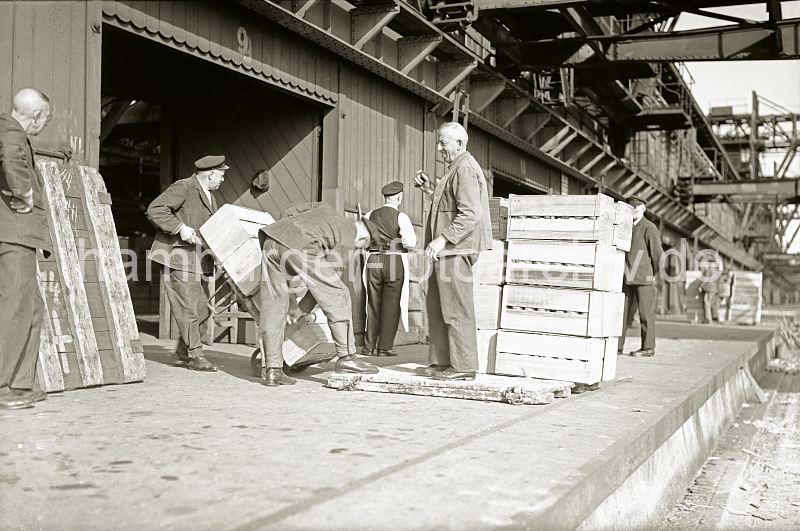 454_6 Der Kran hat eine Hieve Apfelsinenkisten auf der Laderampe eines Lagerschuppens im Hamburger Hafen abgestellt; Hafenarbeiter beladen ihre Sackkarre und transportieren die Sdfrchte an ihren Lagerplatz im Schuppen. Jedes einzelne Tor ist zur Orientierung nummeriert - darber sind die Laufschienen der Halbportalkrane angebracht.  Umschlag von Apfelsinenkisten am Hafenkai - 1934.