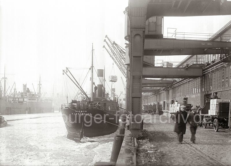 Gterumschlag am Hafenkai - Hieve Apfelsinenkisten am Halbportalkran;  ca. 1930. 454_415 Die Ladung Apfelsinen wird vom Frachter RIGMOR gelscht - Halbportalkrane hieven die Kisten mit den Sdfrchten aus dem Laderaum des Frachtschiffs und stellen die Hieve oben auf der Galerie des Fruchtschuppens  ab. Dort warten die Lagerarbeiter auf die Ladung, um die Fruchtkisten im Lagerschuppen zu verstauen. An der Laderampe stehen Pferdefuhrwerke, die mit Apfelsinenkisten beladen sind.