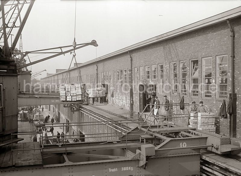 Hieve Apfelsinenkisten - Galerie Fruchtschuppen;  ca. 1930. 455_416 Die Hafenkrane bringen die Hieve Apfelsinenkisten zur Galerie des Fruchtschuppens - dort warten die Lagerarbeiter auf die Ladung, um die Fruchtkisten im Lagerschuppen zu verstauen. Die Halbportalkrane bewegen sich auf Schienen an der Front des Schuppens. Unten auf dem Kai stehen mit Apfelsinenkisten beladene Pferdefuhrwerke an der Laderampe.