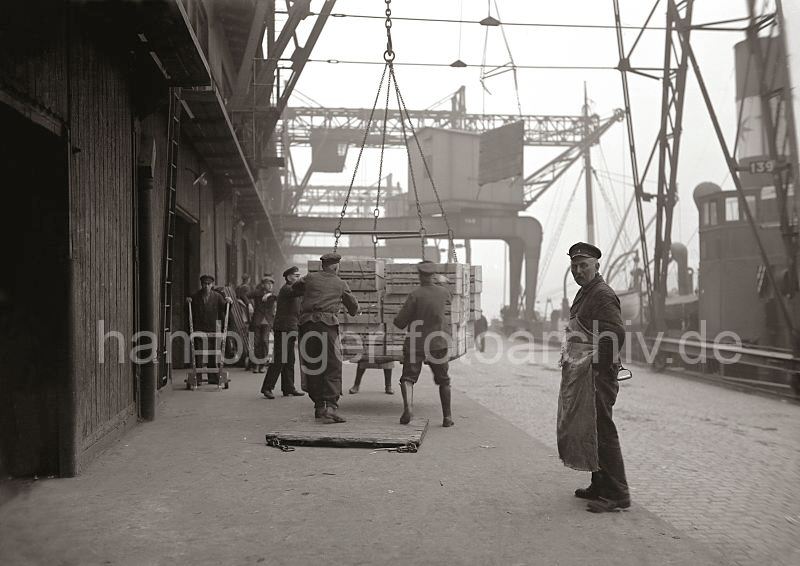 Alte Hamburg Fotografien aus dem Hafen Hamburgs - Kunstdrucke auf Fotoleinwand / Posterprint als Geschenk fuer Buero und Wohnung. Ladung lschen, Apfelsinenkisten - Hafenarbeiter Laderampe Fruchtschuppen;  ca. 1930.  456_153 Die Hieve Apfelsinenkisten wird von den Lagerarbeitern auf der Laderampe des Fruchtschuppens in Empfang genommen. Weitere Hafenarbeiter warten mit ihren Sackkarren darauf, die Kisten mit den Sdfrchten ins Schuppeninnere zu bringen. Die leere Holzplatte mit den sen an den Ecken wird danach wieder mit dem Kran an Bord des Frachtschiffes gebracht - im Hintergrund schwebt eine der Bodenplatten am Ladegeschirr ber der Kaianlage Richtung Schiff.