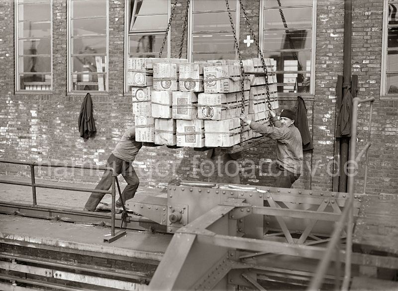 458_417 Ein Kranfhrer setzt die Hieve Apfelsinenkisten auf der Galerie des Fruchtschuppens ab - Lagerarbeiter dirigieren die Ladung an ihren Platz; im Vordergrund das Schienensystem mit Fhrung des Halbportalkrans.  Historische Arbeitsfotos aus dem Archiv der Hamburger Hafen und Logistik AG. Alte Bilder aus dem Hamburger Hafen - Hamburgmotive auf Fotoleinwand / Posterprint als Geschenk fuer Buero und Wohnung.