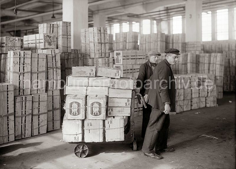 460_419 Zwei Lagerarbeiter transportieren Apfelsinenkisten mit einer Karre im Lager des Fruchtschuppens im Hamburger Hafen. Im Hintergrund sind die Kisten mit den Sdfrchten gestapelt.  Historische Hamburgbilder + Hamburgensien auf Fotoleinwand und Posterprint; als Geschenk fuer Buero und Wohnung.