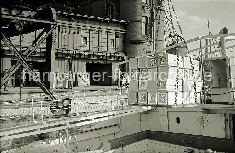 Historische Hamburgbilder + Hamburgensien auf Fotoleinwand und Posterprint; als Geschenk fuer Buero und Wohnung. Ladung lschen eines Frachtschiffs - Apfelsinenkisten am HafenKran. 461_OF_46A Eine Hieve Apfelsinenkisten hngt am Ladegeschirr ber dem Laderaum des Schiffs - auf dem Kai das Laufrad und die Schiene des fahrbaren Halbportalkrans vor dem Fruchtschuppen. Auf der Laderampe stehen die Hafenarbeiter und warten darauf, die Ladung in Empfang zu nehmen.