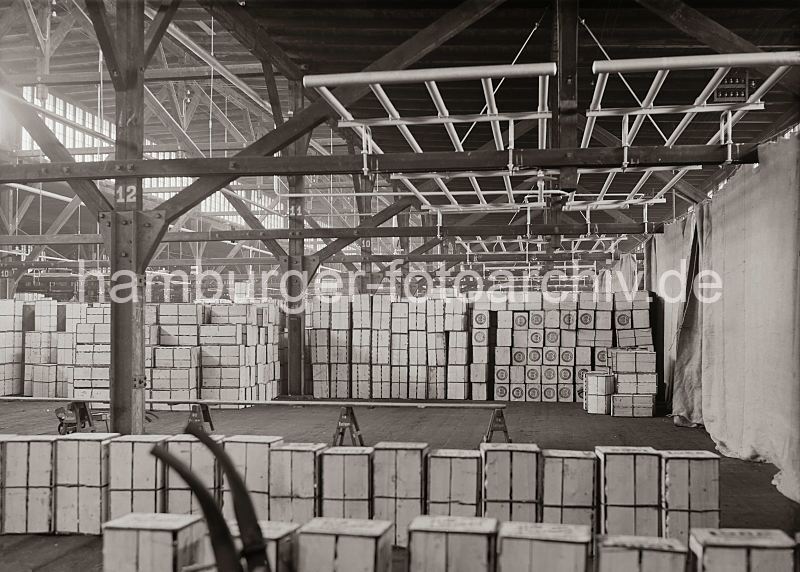 Lagerung von Apfelsinenkisten im Hafen Fruchtschuppen - Hallenkonstruktion. Historische Hamburg Bilder als Geschenk fuer Buero und Wohnung. 464_644a Die Apfelsinenkisten stehen gestapelt in dem klimatisierten Fruchtschuppen - im Vordergrund die Griffe einer Sackkarre. Die Wand ist mit Planen abgehngt, die vor Temperaturschwankungen schtzen soll. Die Holzstnder der Balkenkonstruktion sind mit Nummern zur Bestimmung des Lagerplatzes versehen. Fenster unter dem Dach des hohen Lagergebudes sorgen fr ausreichendes Licht.