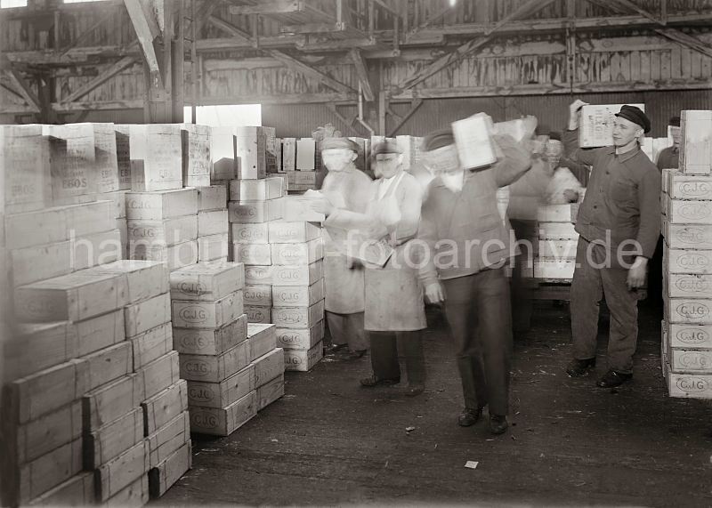 historische Arbeitsbilder aus dem Hamburger Hafen - Kisten stapeln im Fruchtschuppen; ca. 1932. Bilder von der Geschichte Hamburgs - Arbeit im Hafen Hamburg.  471_577 Die gelschten Rosinenkisten sind am Hafenkai auf einen Transportwagen geladen worden - im Fruchtschuppen werden die Kisten von Hand abgeladen und von den Lagerarbeitern auf der Schulter zu dem Lagerplatz gebracht.