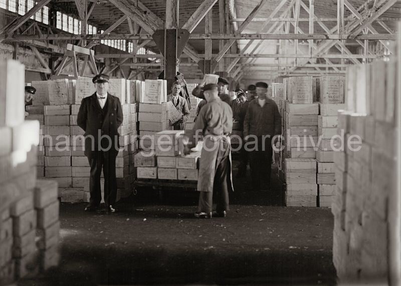 historische Fotografien aus der Arbeitswelt im Hamburger Hafen, Arbeit im Fruchtschuppen; ca. 1932. Bilder von der Geschichte Hamburgs - Arbeit im Hafen Hamburg. Alte Hafenbilder aus dem Archiv der Hamburger Hafen und Logistik AG 472_577a Die Lagerarbeiter nehmen die Kisten mit den Rosinen von dem Transportkarren auf und bringen sie auf der Schulter zur dem Lagerplatz. Die Kisten gefllt mit getrockneten Weinbeeren sind in dem Fruchtschuppen mannshoch aufgestapelt.
