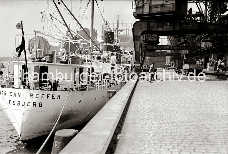 Historische Fotografien aus dem Bildarchiv der HHLA. Heck des Frachters AFRICAN REEFER - Kaianlage vor dem Fruchtschuppen A; ca. 1932.  477_14_B_265a Der dnische Frachter AFRICAN REEFER mit Heimathafen Esbjerg liegt am Versmannkai vor dem Fruchtschuppen A. Seine Ladung wird mit den Halbportalkranen gelscht und der Rampe des Schuppens abgesetzt. Dort nehmen Arbeiter die Fruchtkisten entgegen und transportieren diese mit Sackkarren in das Innere des Lagers. Im Hintergrund das Gasometer der Gasanstalt auf dem Grasbrook.