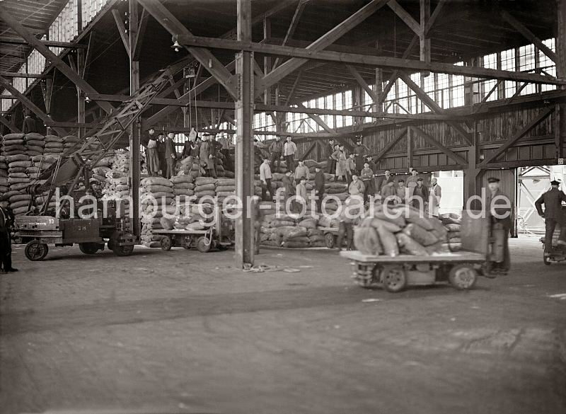 Fotos zur Geschichte des Hafen Hamburgs - Elektrokarren + Schuppenlaufkran; Hafenarbeiter auf gestapelten Scken. 487_409 Das Sackgut wird von Hand im Lagerschuppen gestapelt - Elektrokarren bringen die Scke von der Laderampe, die Arbeiter schichten die Jutescke im Treppenstapel auf. Ein wendiger Schuppenlaufkran mit drei Rdern untersttzt die Lagerkrfte bei ihrer harten Arbeit.