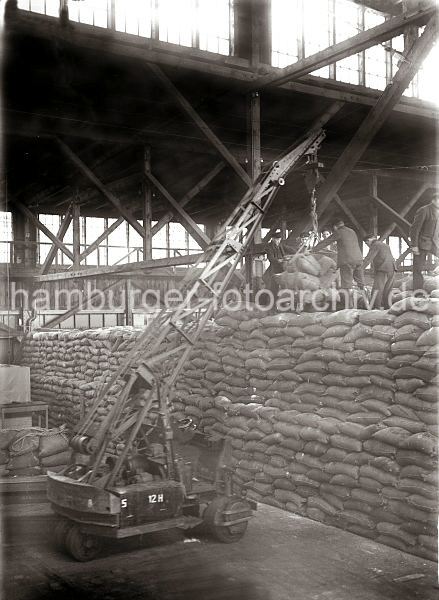 Bilder aus dem Fotoarchiv der Hamburger Hafen und Logistik AG HHLA. Schuppenlaufkran im Kaischuppen Hafen Hamburg; ca. 1934. 491_061_80 Ein beweglicher Schuppenkran hievt eine Ladung Scke zu den Arbeitern auf dem hohen Sackstapel. Die Lagerarbeiter lsen die Hieve vom Kranhaken.