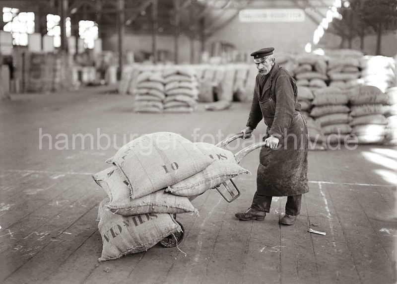 Historische Hafenbilder als Fotoprint, Hochglanz oder Matt - Fotoposter als Geschenk fr Weihnachten, Geburtstag, Jubilaeum oder zur Dekoration. 494_421 MDer Lagerarbeiter hat seine Sackkarre mit vier prall gefllten Jutescke beladen und transportiert seine Ladung im Kaischuppen des Hamburger Hafens. Im Hintergrund ist weiteres Sackgut gelagert.