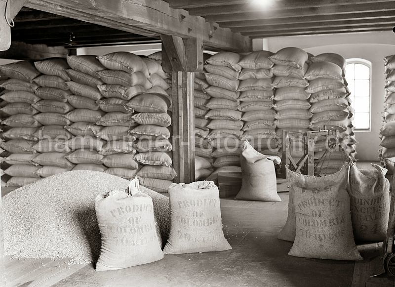 Scke mit Kaffee aus Kolumbien - aufgeschtteter Rohkaffe, Lastwaage - Speicherstadt Hamburg; ca. 1938 - 506_505 Der Kaffee aus Kolumbien ist in Jutescke vernht und bis an die Decke des Speicherbodens gestapelt. Die Scke tragen die Aufschrift Produkt of Colombia - 70 Kilos. Ein Haufen Kaffee ist auf dem Lagerboden ausgeschttet - im Hintergrund eine Waage, mit der die Scke gewogen werden.