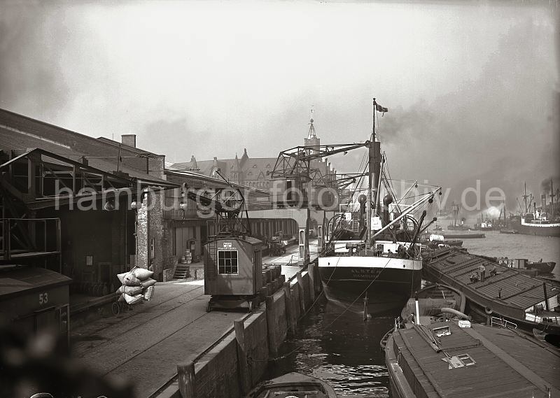 Bilder aus dem alten Hafen Hamburgs - Fotoarchiv der Hamburger Hafen und Logistik AG HHLA - Hafenkrane am Kaiserkai - Sandtorhafen; Ladung lschen - Frachter und  Schuten; ca. 1932. 508_086 Die Ladung eines Frachters wird am Kaiserkai des Sandtorhafens gelscht - im Hintergrund der Turm des Zeitballs vom Kaiserspeicher / Kaispeicher A. Hafenkrne fahren auf Schienen auf der Kaianlage entlang - eine Hieve Scke hngt am Ausleger des Kaikrans. Am hinteren Lagerschuppen arbeiten Halbportalkrane unter deren Auslegern die Lastwagen und Gterzge hindurch fahren knnen. Ein Frachtschiff hat am Kai fest gemacht - seine Ladung wird auch wasserseitig auf Schuten und Binnenschiffe gelscht.