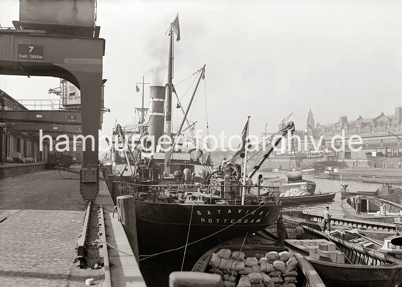 Alte Fotos aus dem Hafen Hamburgs - Bildarchiv der Hamburger Hafen und Logistik AG (HHLA) - Halbportalkrane im Sandtorhafen / Kaiserkai - Ladegeschirr Frachtschiff. 509_721a Ein Frachter aus Rotterdam liegt am Kaiserkai; der Sandtorhafen wurde 1866 als erstes knstliches Hamburger Hafenbecken gebaut, in dem die Frachtschiffe ber die Kaianlage abgefertigt wurden. Das Frachtschiff hievt mit bordeigenem Ladegeschirr eine Tonne von einem Lastkahn an Bord. Weitere Schuten beladen mit Scken und Kisten liegen lngsseits des Schiffs. Im Hintergrund der Sandtorkai und die neogotischen Backsteingiebel der Speicherstadt.
