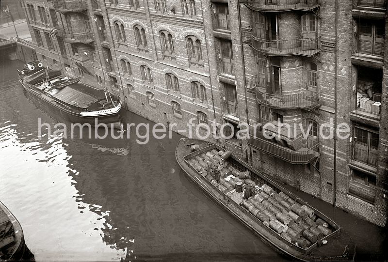 Alte Hamburgbilder aus dem Hafen und der Speicherstadt - Fotoarchiv der HHLA - Fleet in der Speicherstadt, Hamburger Freihafen - eine Ladung Fsser wird gelscht.  515_B_342a Ein Motorschiff und eine Schute liegen an der Mauer eines Lagerschuppens auf einem Fleet in der Hamburger Speicherstadt. Die Ladung der Schiffe wird ber Winden gelscht. Eine Hieve Scke wird zum Lagerraum hochgezogen und ein Fass hngt an dem Kranhaken ber des Laderaum der Schute. Durch eine geffnete Tr sind Kisten und Scke auf einem Lagerboden zu erkennen.