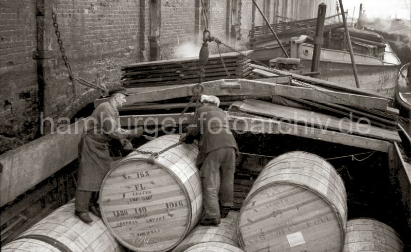 Alte Hamburgfotografien aus dem Hafen und der Speicherstadt der HHLA Tabakfsser im Saalehafen - Lagerhaus G, Arbeiter legen das Ladegeschirr an; 1948.  516_0001 Die grossen Holzfsser mit Tabak sind im Laderaum der Schute gestapelt. Zwei Arbeiter legen das Ladegeschirr an dem Fass an, damit es in den Laderaum des Lagerhaus G am Saalehafen gehoben werden kann. Im Hintergrund liegen weitere Schuten an der Mauer des Ziegelgebudes.