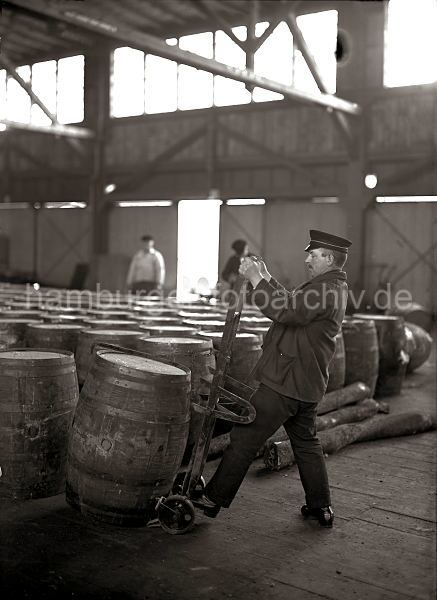 Geschichte des Hamburger Hafens - Bilder aus dem Fotoarchiv der HHLA Selbstladekarre mit Fass - Lagerarbeiter im Kaischuppen; ca. 1930.  520_68 Ein Arbeiter setzt eine Holztonne auf die Selbstladekarre; mit Hilfe eines Eisenhakens wird das Fass auf die Sackkarre gezogen.