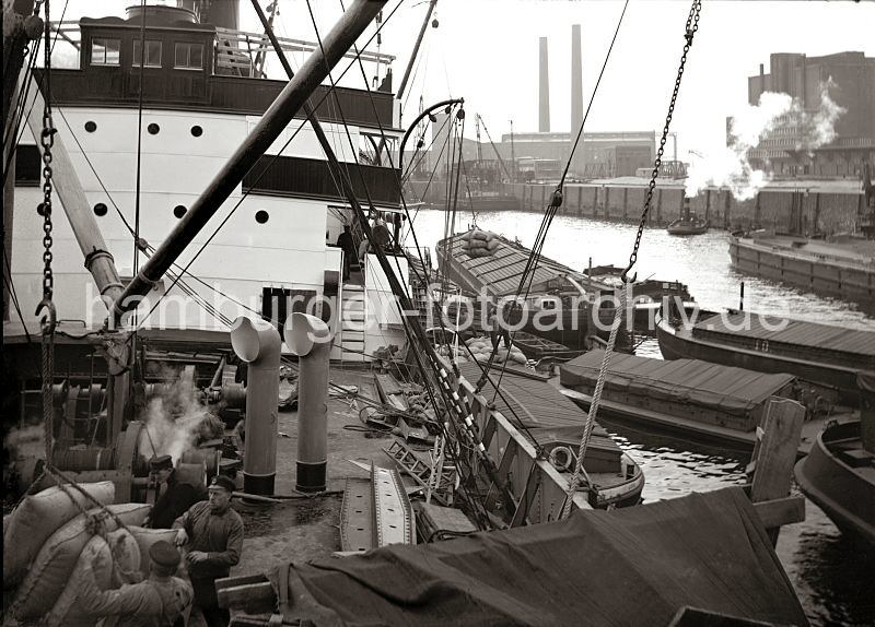 Historische Bilder vom Gterumschlag im Hafen Hamburg / Bildarchiv vom Hamburger Hafen. Die Ladung eines Frachters wird gelscht - Hieve mit Scken am Kranhaken - Kraftwerk HEW + Khlhaus fr Eier. 535_151 Ein Frachtschiffs nimmt im Oderhafen Ladung auf - ber bordeigene Ladebume werden Scke von den lngsseits liegenden Schuten und Lastkhne gehievt. Im Vordergrund nehmen zwei Schauerleute die Hieve Scke, die an einem Kranhaken hngt entgegen. Im Hintergrund die Schornsteine des HEW Elektrizittswerks an der Nippoldstrasse / Freihafenstrasse. Rechts der Khlspeicher fr importierte Eier am Ro Kai des Ro Hafens.