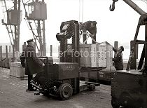 543_184 Eine große Holzkiste ist mit einem Elektrohubwagen der AEG von seinem Fahrer auf die Rampe des Schuppen 83 am Chilekai des Hamburger Oderhafens transportiert worden. Hafenarbeiter legen Tauwerk um die Kiste, an dem der fahrbare Kran sie auf den bereitstehenden Güterwaggon heben wird.