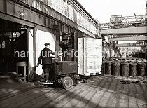 546_143 Ein Lagerarbeiter hat eine große Holzkiste auf die Gabel des Elektrohubwagen geladen und fährt sein Transportgut auf die Lagerrampe des Schuppens 83 am Chilekai des Oderhafens. Auf der Rampe stehen Metallfässer, im Hintergrund ein Arbeiter mit einer Sackkarre.