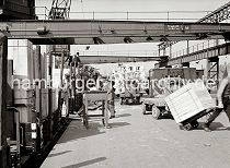 549_391 Ein Güterzug wird an der Laderampe entladen, er hat Stühle und Kisten geladen. Ein Arbeiter transportiert eine grosse Holzkiste mit einer Sackkarre, ein anderer Hafenarbeiter fährt gerade mit der Karre die Laderampe auf den Güterwaggon hoch. Im Hintergrund ein Elektrokarren, der Stühle aufgeladen hat.
