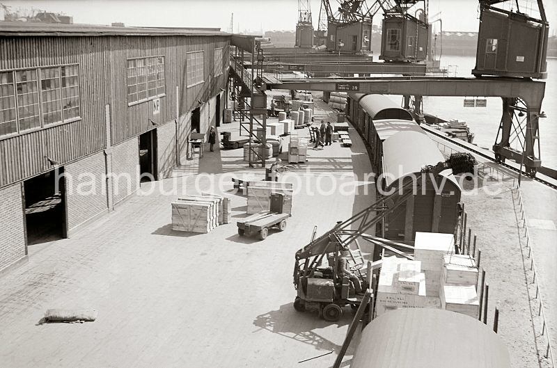 Historische Bilddokumente aus dem Hafen Hamburgs / Blick aus dem Fhrerhaus des Hafenkrans - Gterzug an der Laderampe. Hamburg Bilder zu Weihnachten als Geschenk! 556_394a Blick von einem Hafenkran auf das geschftige Treiben im Hamburger Hafen. Kisten stehen zum Abtransport bereit - ein fahrbarer Schuppenkran hat gerade eine Holzkiste auf dem offenen Gterwaggon abgesetzt. Im Hintergrund werden Kisten und Ballen auf die Anhnger der Elektrokarren geladen.