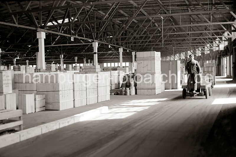 <title>Historische Fotodokumente aus dem Hafen Hamburgs. Elektrokarren mit Transportarbeiter im Hamburger Hafen - Kistenlagerung im Lagerschuppen; Firma Jubilaeum - Geschenk Bilder Hamburg. 557_390a Ein Transportarbeiter fhrt seine Ladung mit dem Elektrokarren und ihren Anhngern in den Lagerschuppen des Hamburger Hafens. Dort sind unterschiedlich grosse Holz-Kisten gestapelt, die auf ihren Weitertransport warten. 