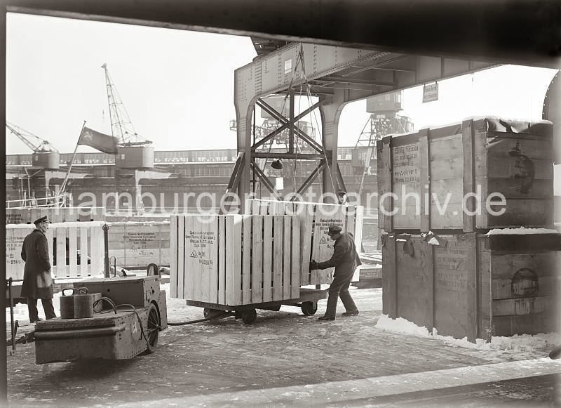 Bilddokumente aus dem Hafen Hamburgs. Elektrokarren mit Anhnger, Verladung Holzkiste am Kaischuppen - Hafenarbeiter bei der Arbeit; Geschenk fure eine Firma zur Gruendung in Hamburg.  558_522 Eine groe Holzkiste ist von einem sowjetischen Frachter gelscht worden, dessen Flagge mit Hammer und Sichel am Heck des Frachtschiffs im Hintergrund weht.  Die Kiste hngt am Ladegeschirr des Halbportalkrans und wird gerade von einem Hafenarbeiter auf den Anhnger eines Elektrokarrens abgesetzt.