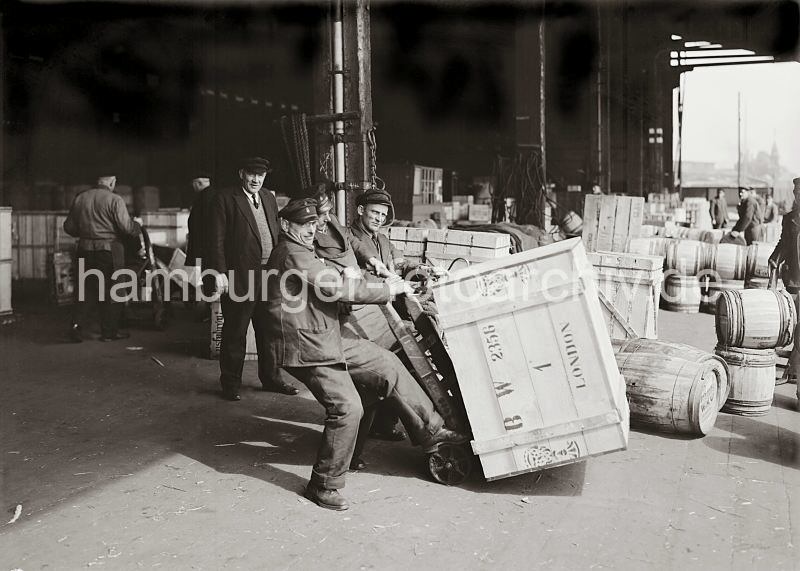 Alte Hafenfotos aus Hamburg - Arbeitsbilder. Kistentransport mit Sackkarre - Lagerarbeiter im Kaischuppen - Hamburg-Bilder als Geschenk zur Einladung Jubilaeumsfeier. 559_596a Drei Lagerarbeiter stemmen eine schwere Holzkiste auf eine Sackkarre. Im Hintergrund stehen weitere Kisten und Holzfsser im Kaischuppen und auf der Laderampe. Arbeiter arbeiten mit Handkarren im Lagerschuppen. 