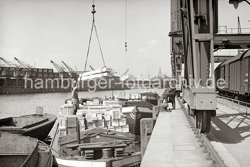 Alte Hamburgfotos von der Arbeit im Hafen der Hansestadt Hamburg; Bildrchiv der HHLA. Gterumschlag am Hafenkai - mit Kisten beladene Schute - Holzkisten am Kranhaken // Fotos als Geschenk zur Einweihung der Firma. 565_395 Ein Lastkahn liegt mit Holzkisten hoch beladen am Hafenkai - eine Hieve Kisten hngt am Kranhaken ber dem Laderaum der Schute. Unter dem Halbportalkran stehen geschlossene Gterzge der Hafenbahn. Im Vordergrund Rder und Laufschiene der Krananlage.