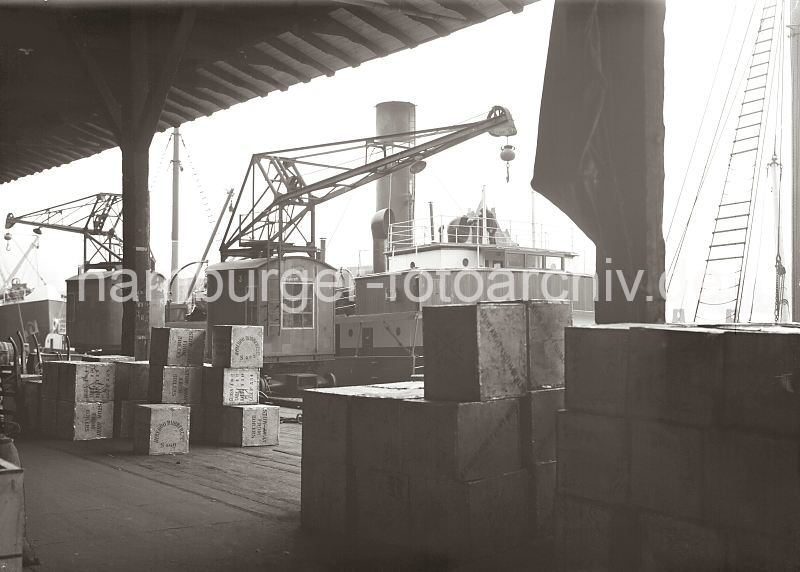 Hamburg-Fotos als Abschiedsgeschenk fuer Mitarbeiter der Firma. Historische Hafenbilder aus dem Archiv der Hamburger Hafen und Logistik AG, HHLA. Kaikrane -  Frachtschiff, gelagerte Kisten im Hafenschuppen. 568_653a Hafenkrane an einem Kai des Hamburger Hafens - ein Frachtschiff hat an der Kaianlage fest gemacht, der hohe Schornstein und das Steuerhaus sind zu erkennen. Auf dem Kais stehen Hafenkrane, die in Schienen am Rande des Hafenbeckens entlang fahren knnen. Holzkisten sind im Lagerschuppen gestapelt - lks. im Hintergrund ragen die Griffe von abgestellten Sackkarren aus dem Lagergut heraus.