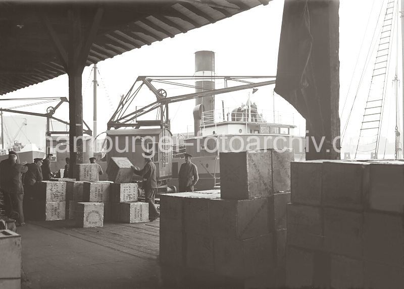 Hamburg Bilder als Praesent zur Einweihung der Firma. Historische Hafenfotografien - Kaikrane lschen die Ladung eines Frachtschiffs, Hafenarbeiter stapeln Kiste. 569_653 Hafenkrane entladen ein Frachtschiff - Hafenarbeiter stapeln die angelandeten Kisten von Hand in dem Lagerschuppen des Hamburger Hafens.  Auf den Kisten ist der Name der Lieferfirma Avreboo Rubber Co. Ltd. aufgedruckt.