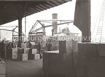 569_653 Hafenkrane entladen ein Frachtschiff - Hafenarbeiter stapeln die angelandeten Kisten von Hand in dem Lagerschuppen des Hamburger Hafens. Auf den Kisten ist der Name der Lieferfirma Avreboo Rubber Co. Ltd. aufgedruckt.