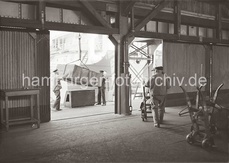 Fotos aus der Geschichte der Hafenarbeit in Hamburg - Entladung eines Frachtschiffs - Holzkiste am Kranhaken; Lagerarbeiter mit Sackkarre; ca. 1934.  574_658a Ein Frachtschiff wird im Hamburger Hafen entladen - eine Holzkiste hngt am Haken des Hafenkrans und wird von zwei Hafenarbeitern auf einem Ladebock abgesetzt. Zwei weitere Lagerarbeiter schieben ihre Sackkarre zur Laderampe; im Vordergrund stehen zwei Sackkarren mit ihren Metall-Laufrdern.