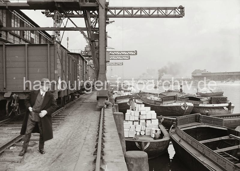 Bilder der Hafengeschichte Hamburgs - Fotos aus dem Archiv der HHLA; Kaianlage   am Afrikakai des Sdwesthafenens - Gterzug der Hafenbahn, Halbportalkrane. 579_645 Schuten liegen am Afrikakai des Hamburger Sdwesthafens - einige der Lastkhne sind leer, whrend ein anderer mit Holzkisten hoch beladen ist. Unter den Halbportalkranen steht ein Gterzug; im Hintergrund dampft ein Schlepper in dem Hafenbecken. Der gegenber liegende Togokai  des ehemaligen Petroleumhafens ist noch nicht fertig gestellt .
